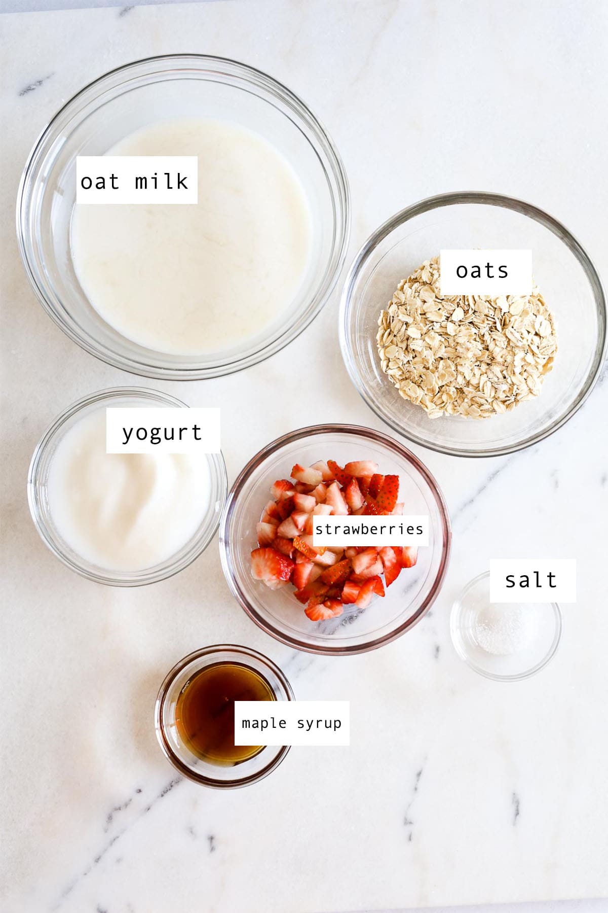 A picture with glass bowls filled with the ingredients to make strawberries and cream oatmeal with a marble background.