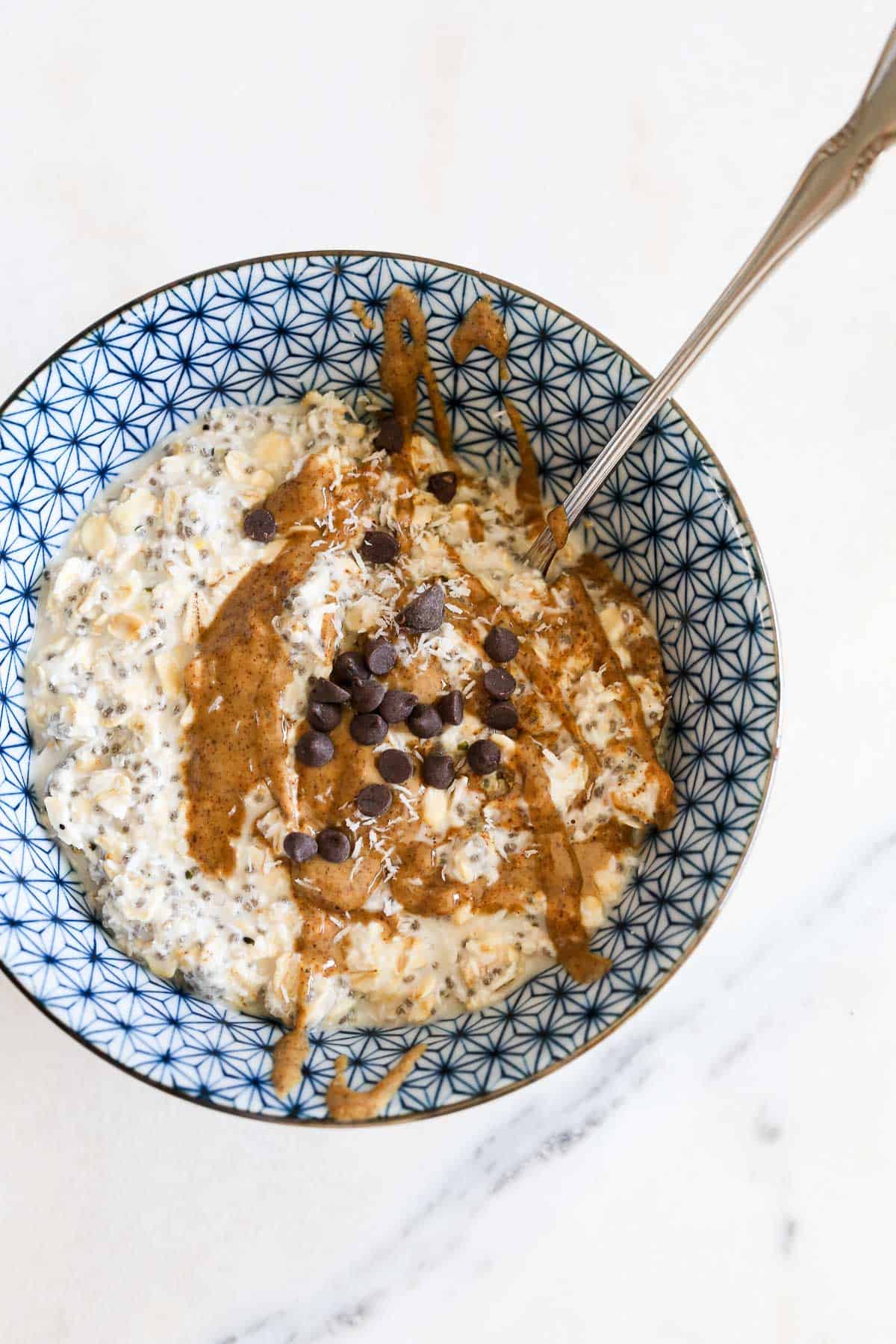 A blue and white printed bowl with coconut overnight oats in it topped with almond butter and chocolate chips sitting on a marble background.